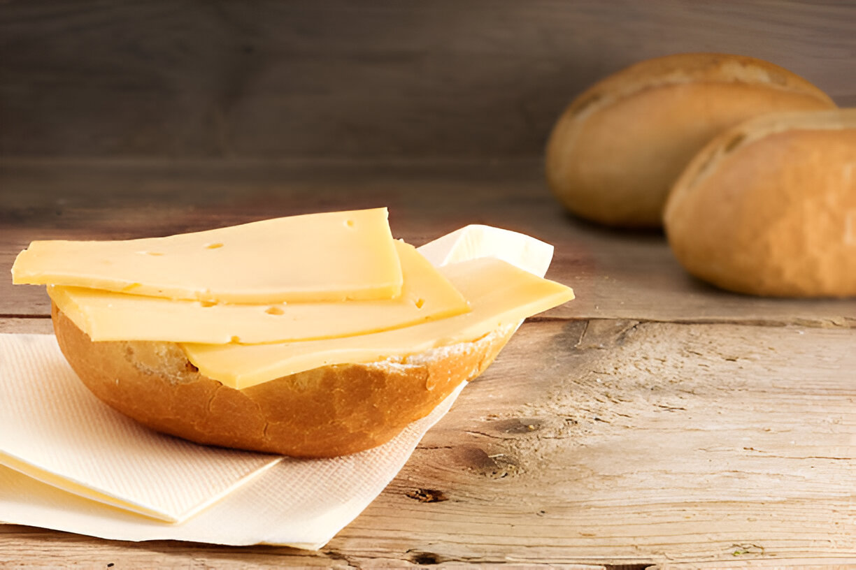 A fresh bread roll topped with slices of yellow cheese on a wooden table, with additional rolls in the background.