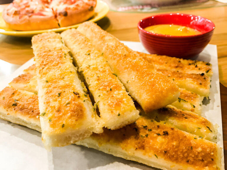 Golden brown garlic breadsticks served with a red bowl of dipping sauce, with a pizza in the background.