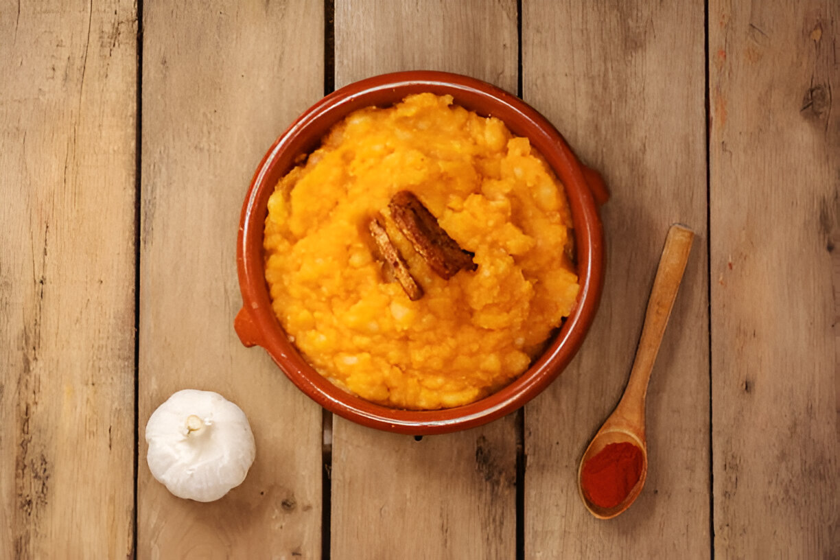 A bowl of mashed orange-colored food with a piece of cinnamon on top, accompanied by a garlic bulb and a wooden spoon with red spice on a wooden table.