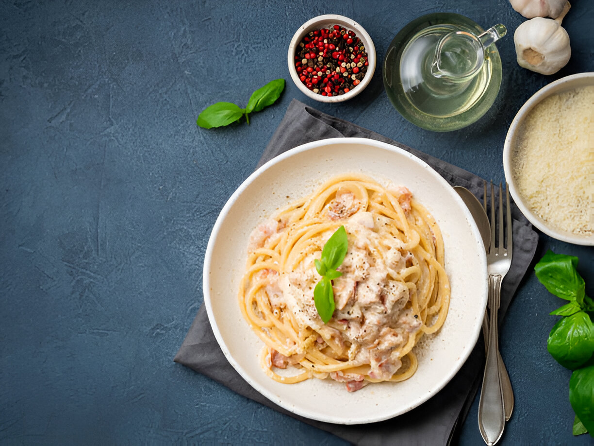 A plate of creamy pasta topped with fresh basil leaves, accompanied by garlic, olive oil, colorful peppercorns, and grated cheese.