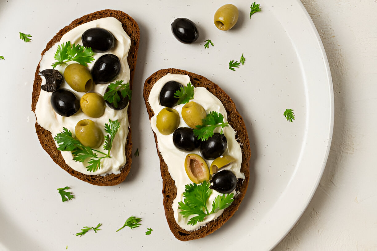 Two slices of dark bread topped with creamy white spread, green and black olives, and garnished with fresh cilantro on a white plate.