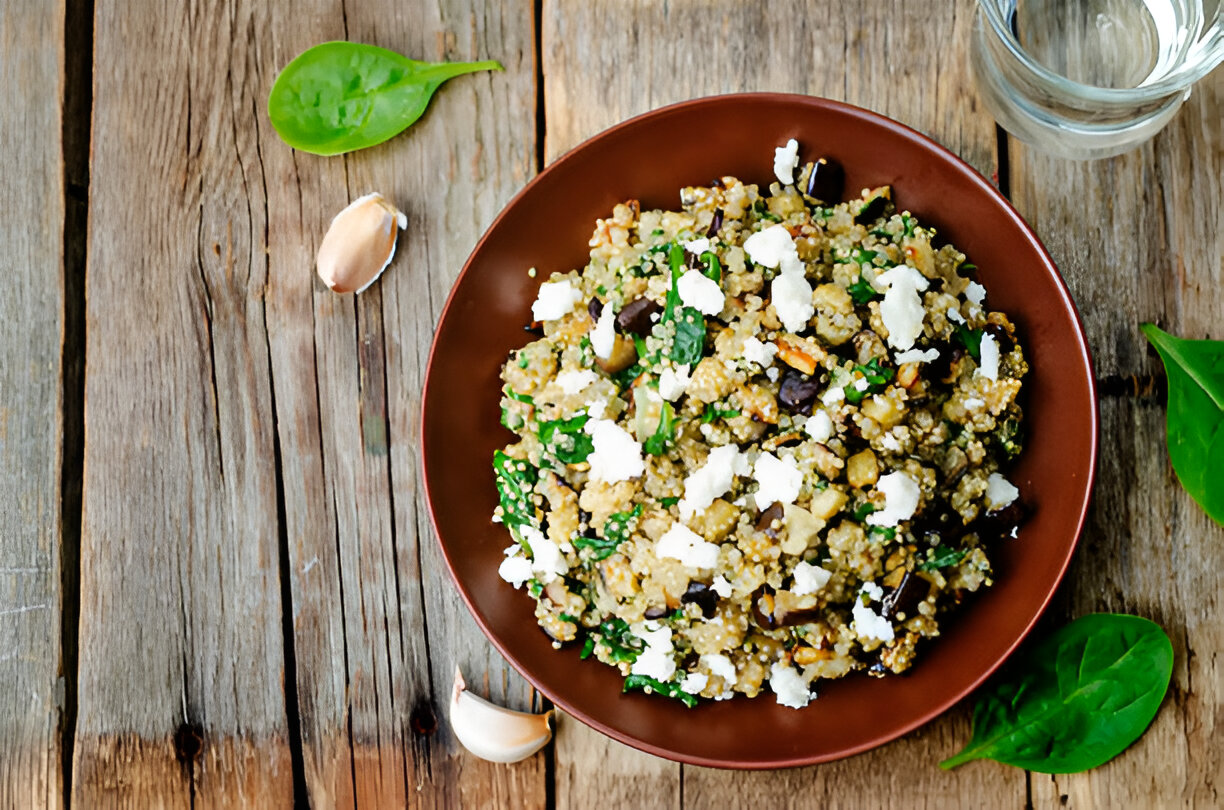 A brown bowl of quinoa salad garnished with spinach, feta cheese, and garlic cloves on a rustic wooden table