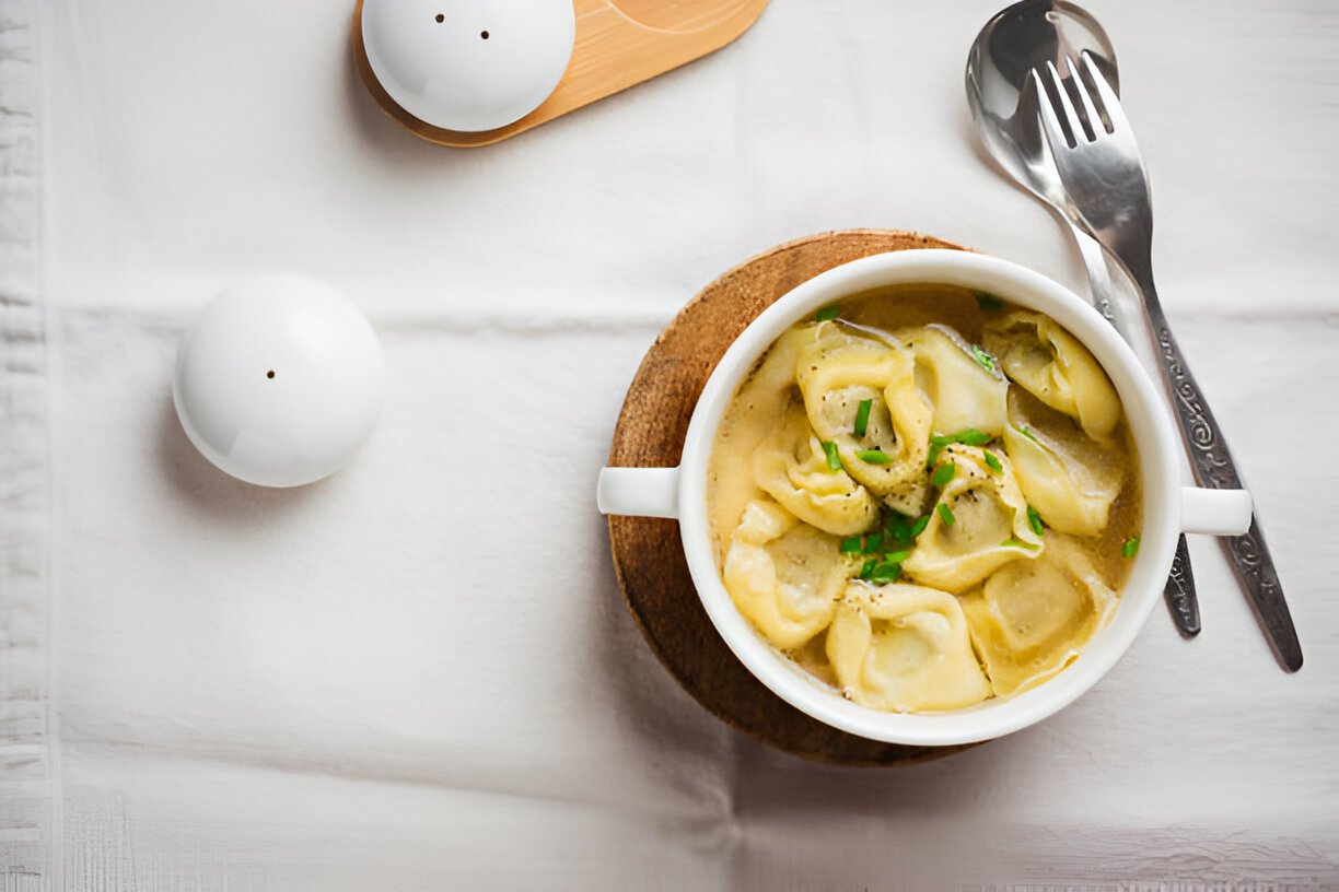 A bowl of dumpling soup with a sprinkle of herbs, accompanied by silver utensils and salt and pepper shakers on a white tablecloth.