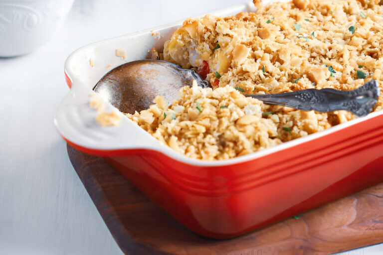 A red casserole dish filled with a baked dish topped with a crunchy breadcrumb crust, a serving spoon resting inside.