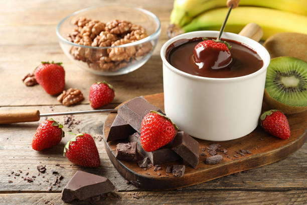 A pot of melted chocolate with strawberries for dipping, surrounded by pieces of dark chocolate, a bowl of walnuts, and fresh fruits.