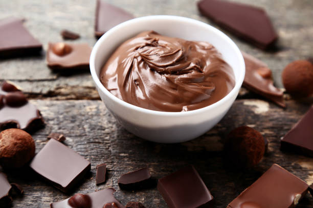 A bowl of creamy chocolate spread surrounded by various chocolate pieces on a wooden surface.
