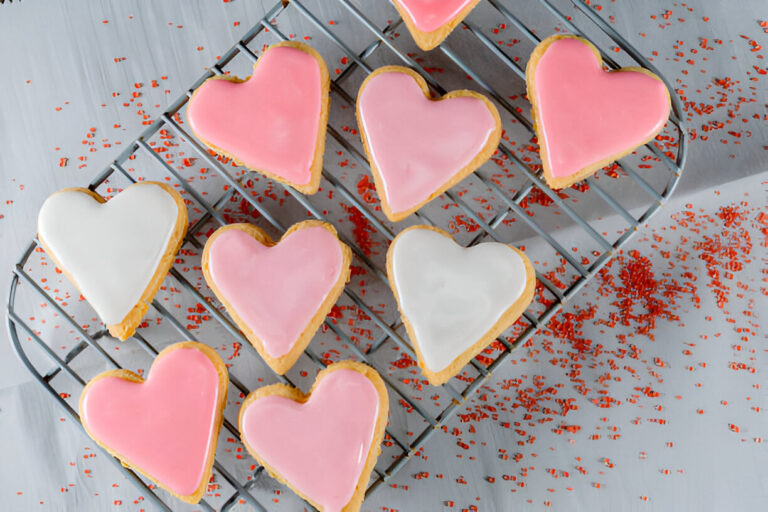 A tray of heart-shaped cookies decorated with pink and white icing, surrounded by red sprinkles.