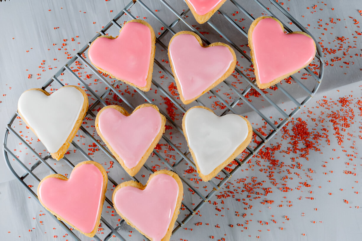 A tray of heart-shaped cookies decorated with pink and white icing, surrounded by red sprinkles.