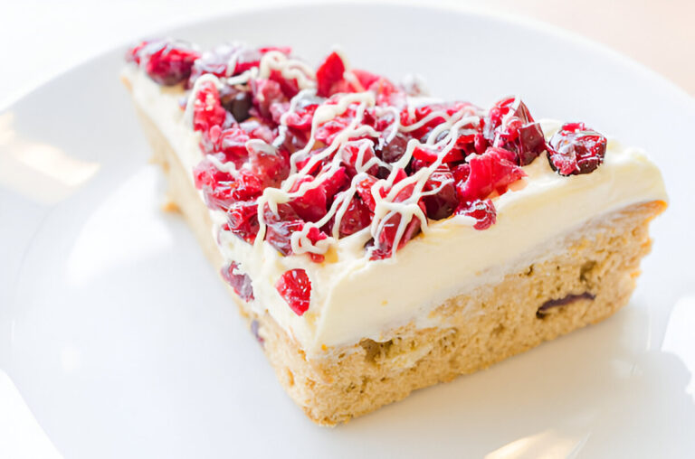 A slice of cheesecake topped with red cranberries and drizzled white chocolate on a white plate.