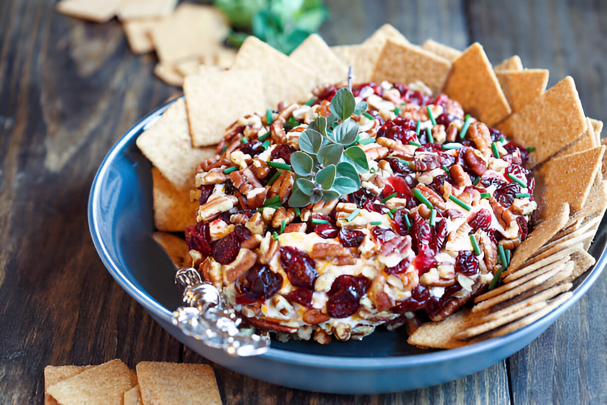 A colorful cheese ball garnished with nuts and herbs, surrounded by graham crackers on a wooden table.