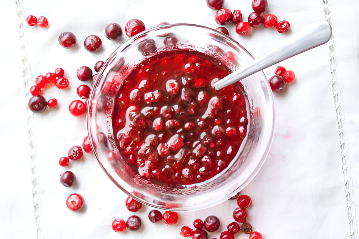 A glass bowl filled with cranberry sauce surrounded by fresh cranberries on a white surface.