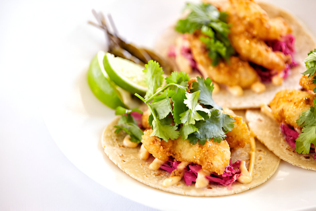 Three tacos topped with crispy fish, cilantro, and pink cabbage, served with lime wedges and jalapeños.