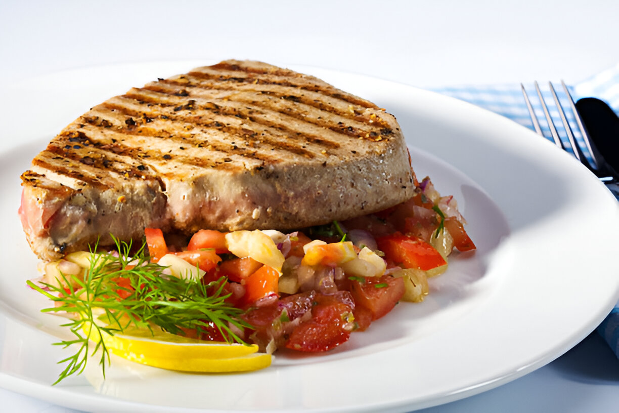 A grilled tuna steak served on a white plate with a fresh tomato salsa and lemon slices.
