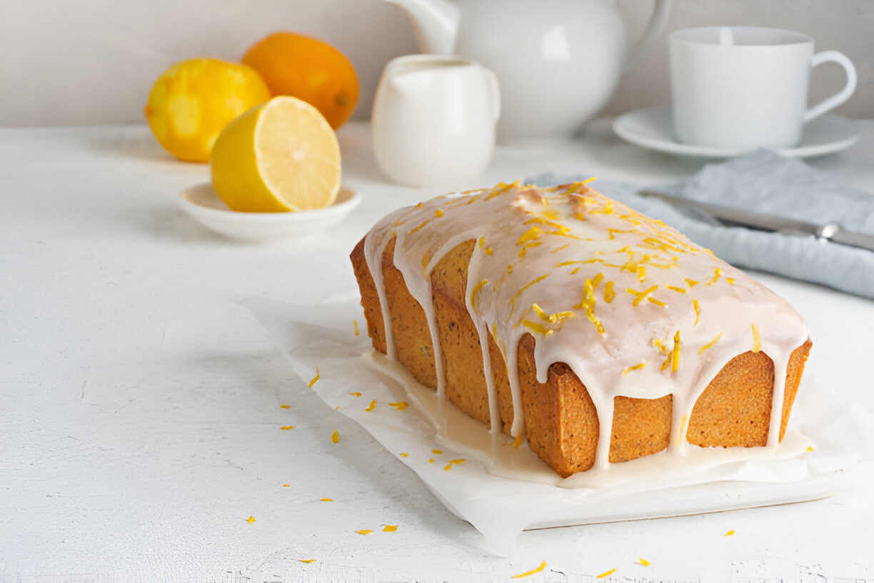 A delicious lemon loaf cake topped with a white glaze and yellow lemon zest, surrounded by fresh lemons and baking accessories.