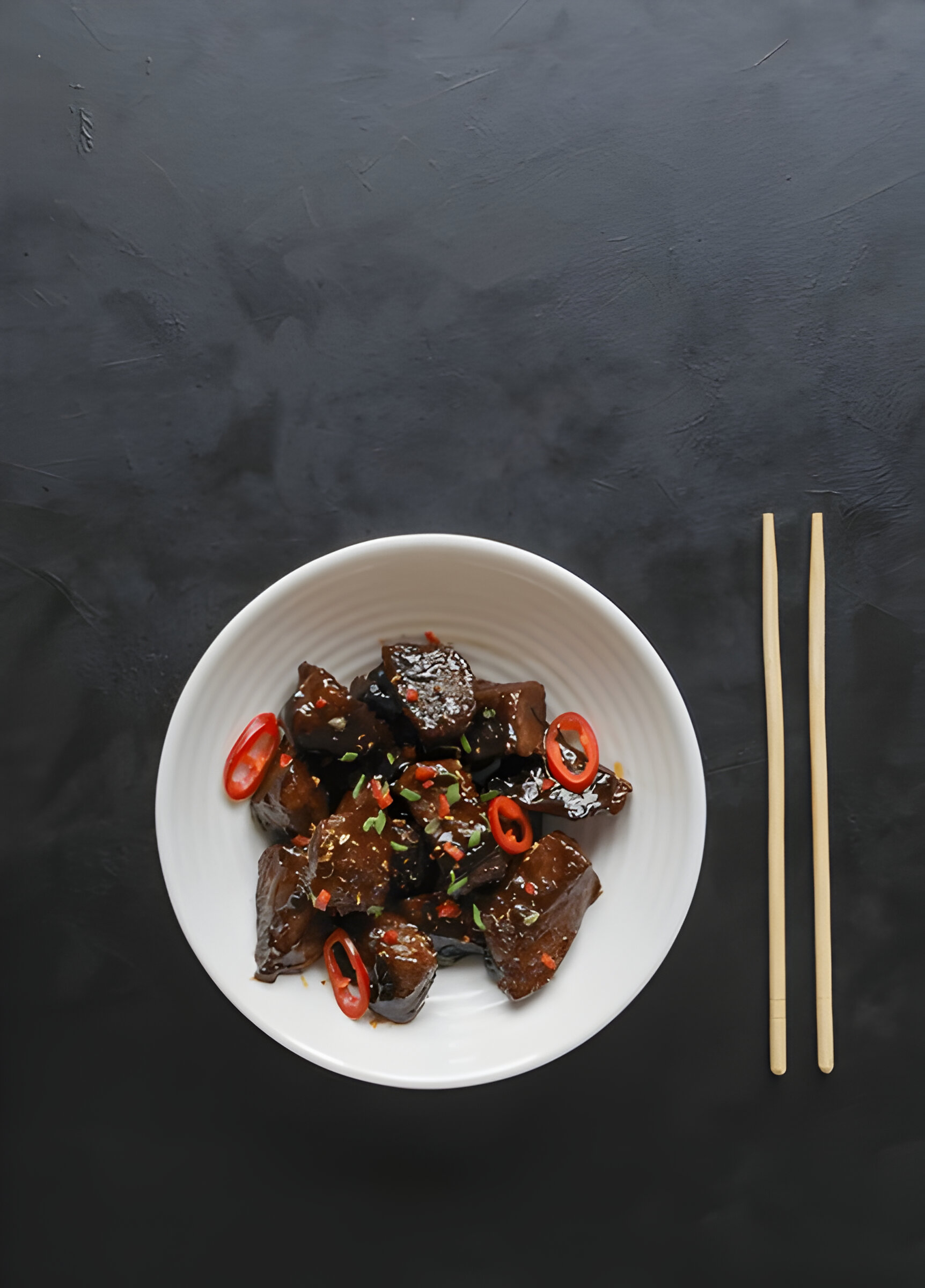 A white bowl of dark, glossy cooked food topped with sliced red chili and herbs, placed on a black surface with wooden chopsticks.