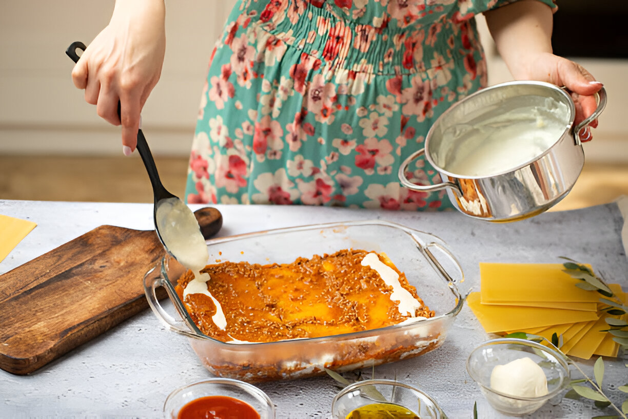 A person in a floral dress prepares a layered dish in a glass baking dish, pouring a creamy sauce over it.