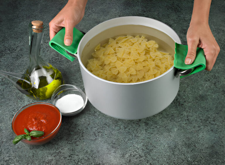 A person is using green silicone handles to hold a pot filled with cooked bowtie pasta. Nearby, there are olive oil, a bowl of tomato sauce, and a small container of salt.