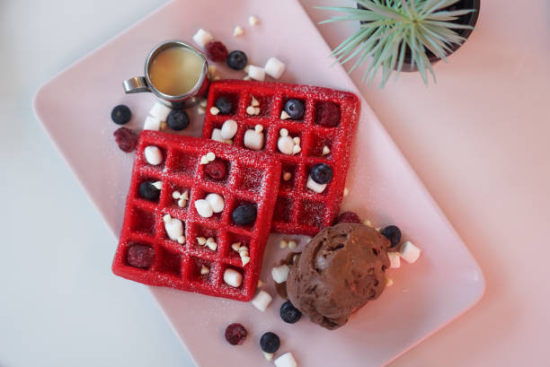 Two bright red waffles topped with chocolate ice cream, blueberries, and marshmallows on a pink plate.