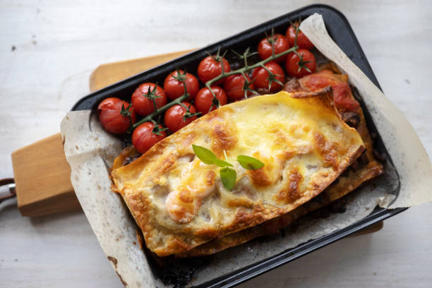 A delicious baked lasagna served in a black tray alongside roasted cherry tomatoes on vine.
