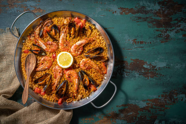 A vibrant seafood paella served in a large pan, garnished with a lemon slice and surrounded by a wooden spoon and burlap fabric on a rustic table.