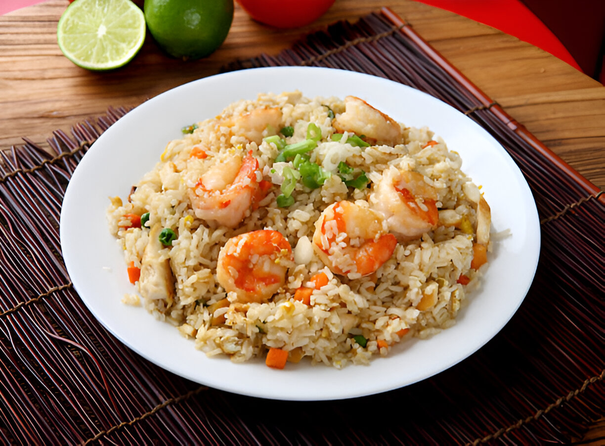 A plate of shrimp fried rice garnished with green onions, served on a bamboo mat with lime and tomato in the background.