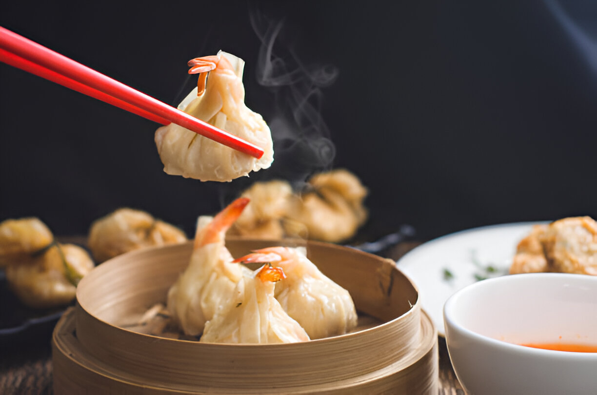 A person using red chopsticks to lift a dumpling with shrimp, steam rising from the dumpling