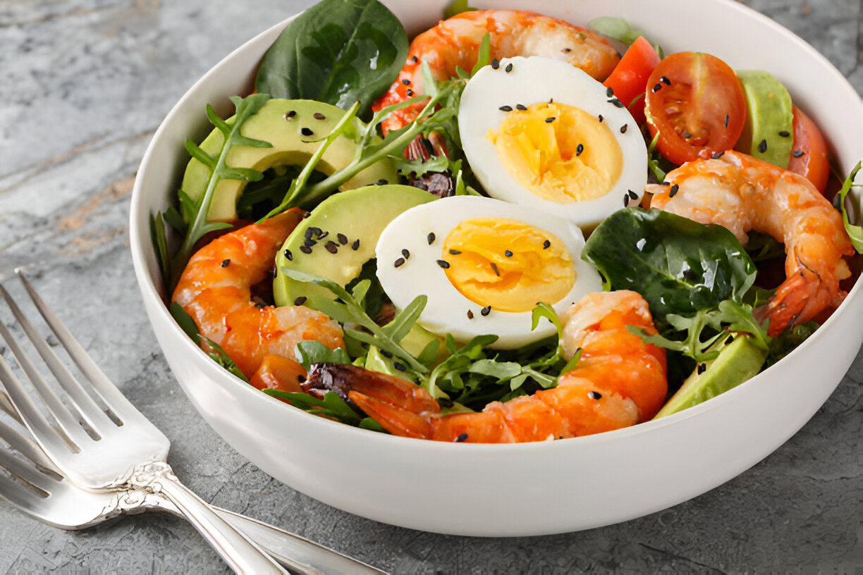 A vibrant shrimp salad featuring fresh greens, avocado slices, cherry tomatoes, and boiled eggs, garnished with sesame seeds.