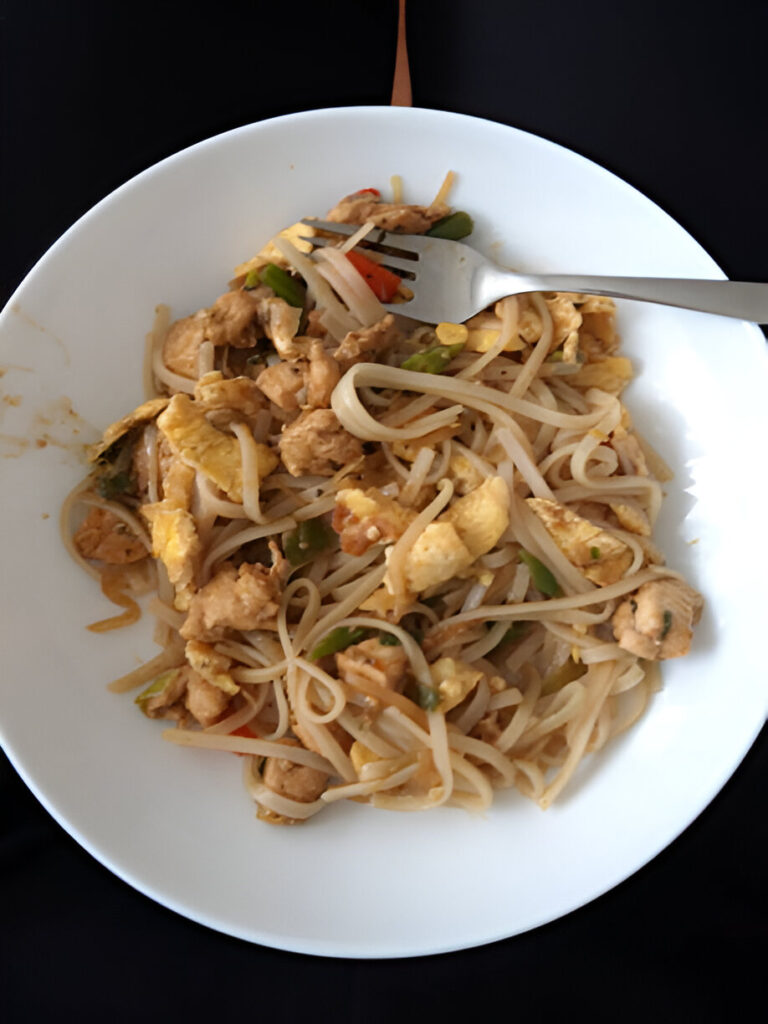 A plate of stir-fried noodles with chicken, vegetables, and egg, garnished with sauce.