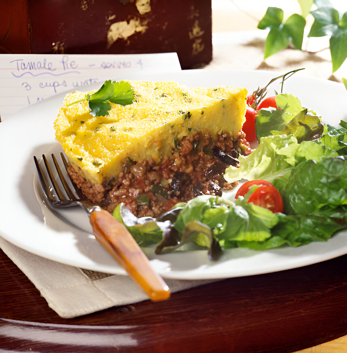 A slice of tamale pie served on a white plate alongside a fresh salad and cherry tomatoes